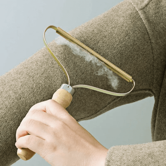 Hand using a manual pet hair remover brush on brown sweater, showcasing its wooden handle and metal frame with lint collection.